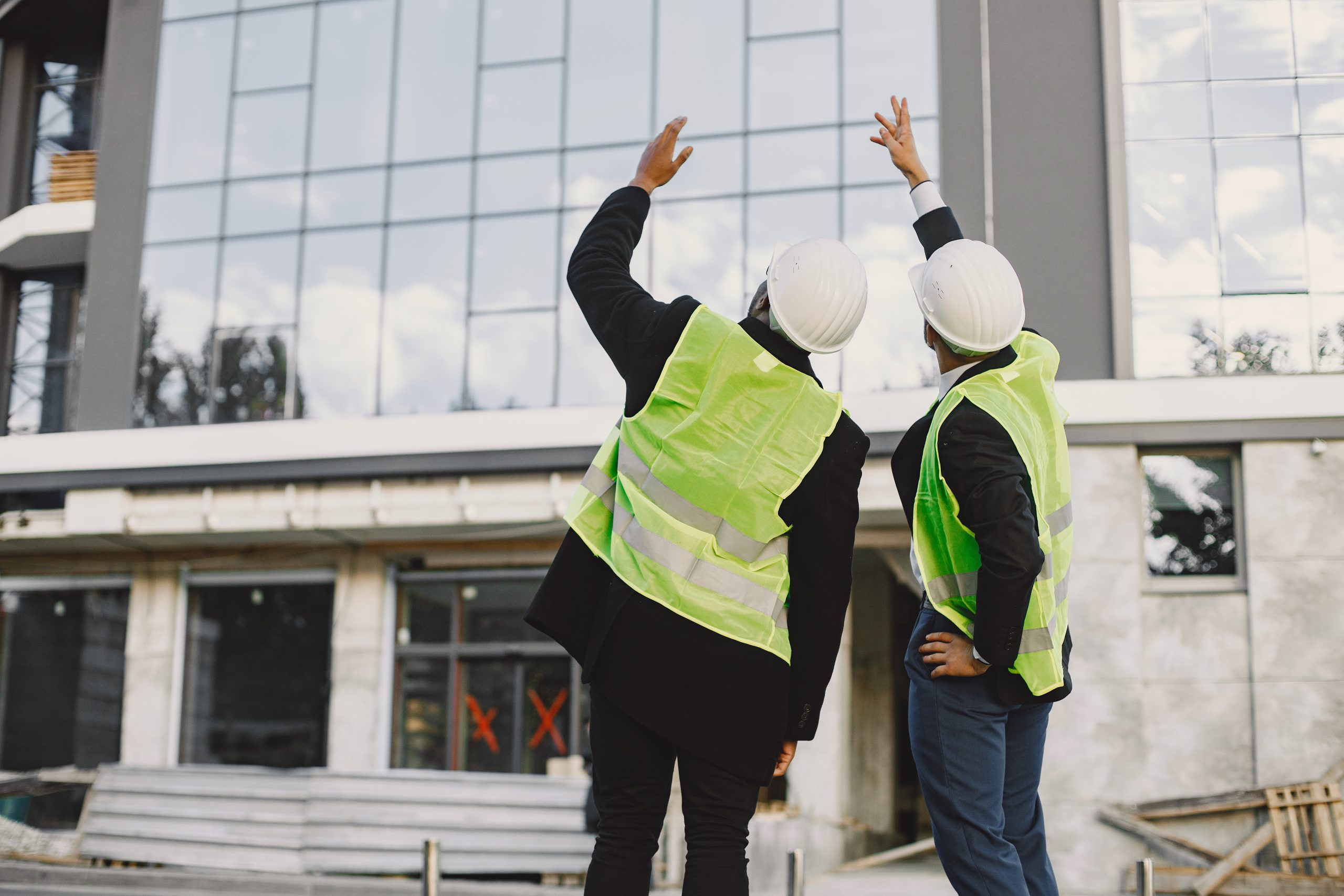 Multi racial builders standing outdoors, back view. Wearing uniform, talking about new glass building. Working on the poject. City infrastructure. Black man.
